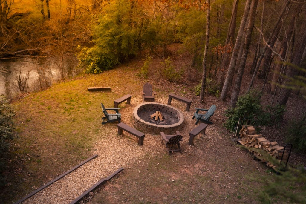 Benches and lawn chairs around a fire pit with firewood stacked on the side towards the back of the Lincoln Logs Cabin. A stream is right beside the fire pit